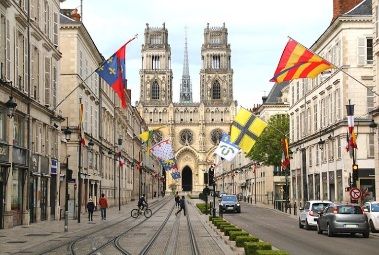 Vue de la cathédrale d’Orléans au fond avec rue bordée de bâtiments, drapeaux colorés et tramway, voitures et piétons. Quartiers à Orléans où habiter, guide.